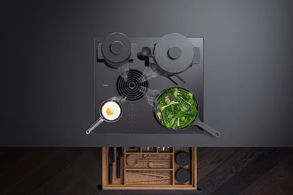 Aufsicht auf schwarzes BORA Kochfeld mit Abzug. Spiegelei und grünes Gemüse braten/dämpfen. Dampf wird abgesaugt. Geöffnete Holzschublade sichtbar.